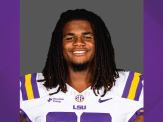 LSU football player smiling in white and purple jersey with LSU logo on gray background.