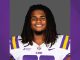 LSU football player smiling in white and purple jersey with LSU logo on gray background.