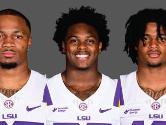 Three LSU football players pose together in white and purple uniforms against a gray backdrop, facing the camera.