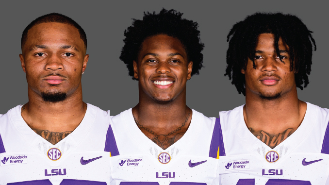 Three LSU football players pose together in white and purple uniforms against a gray backdrop, facing the camera.