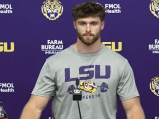 Athlete at a press conference wearing a gray LSU shirt with a microphone in front, backdrop features LSU logos and Farm Bureau branding in purple and gold.