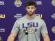 Athlete at a press conference wearing a gray LSU shirt with a microphone in front, backdrop features LSU logos and Farm Bureau branding in purple and gold.
