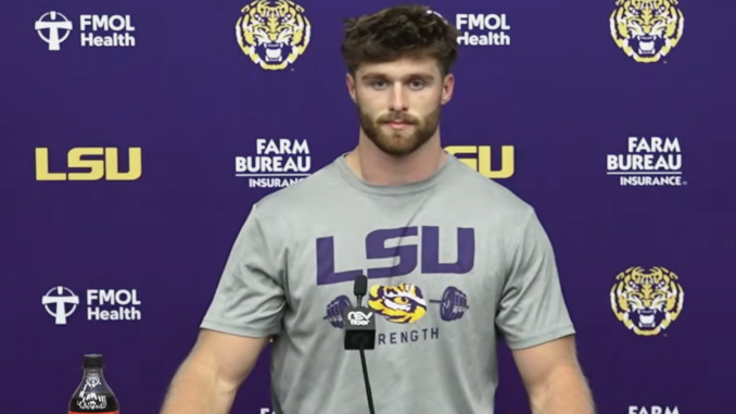 Athlete at a press conference wearing a gray LSU shirt with a microphone in front, backdrop features LSU logos and Farm Bureau branding in purple and gold.