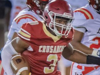 Football player in red Crusaders uniform running with ball during a game, chased by opponents in white uniforms.
