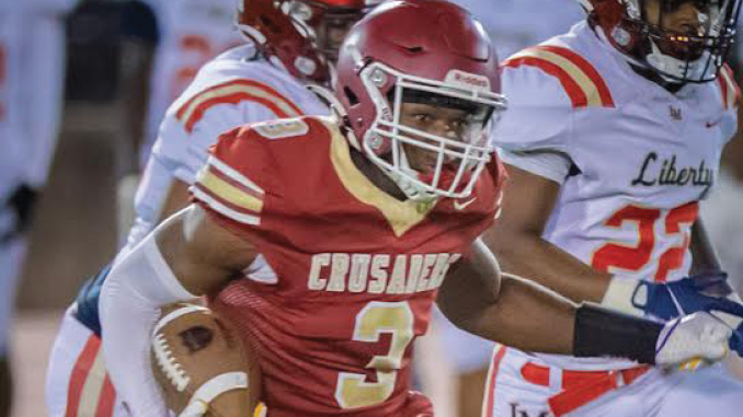 Football player in red Crusaders uniform running with ball during a game, chased by opponents in white uniforms.