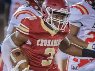 Football player in a red Crusaders jersey with number 3 runs with the ball during a game, pursued by opponents in white uniforms.