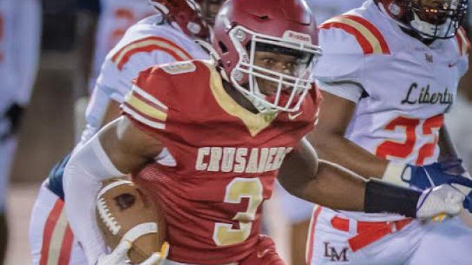 Football player in a red Crusaders jersey with number 3 runs with the ball during a game, pursued by opponents in white uniforms.