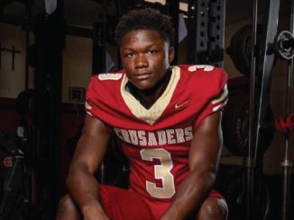 Easton Royal, a Louisiana high school football player in a red jersey with number 3 sits on a weight bench in a gym, looking at the camera.