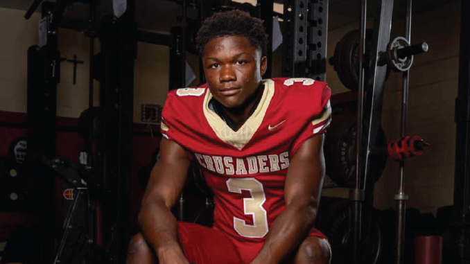 Easton Royal, a Louisiana high school football player in a red jersey with number 3 sits on a weight bench in a gym, looking at the camera.