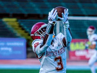Football players on a stadium field as a Brother Martin player (number 3) reaches up to catch a football while a defender in a red helmet closes in behind him.