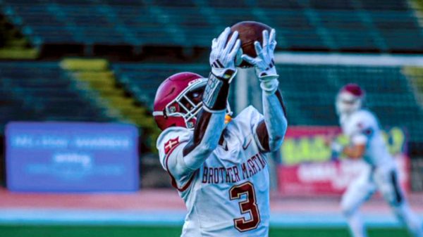Football players on a stadium field as a Brother Martin player (number 3) reaches up to catch a football while a defender in a red helmet closes in behind him.