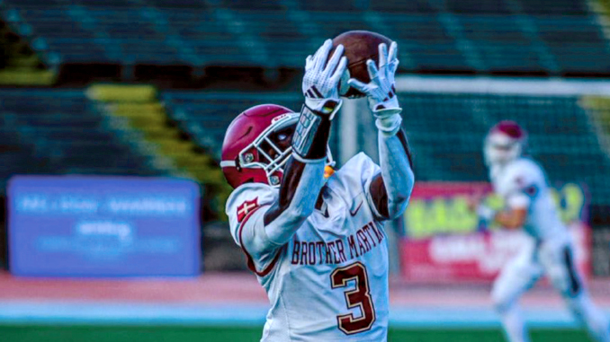 Football players on a stadium field as a Brother Martin player (number 3) reaches up to catch a football while a defender in a red helmet closes in behind him.
