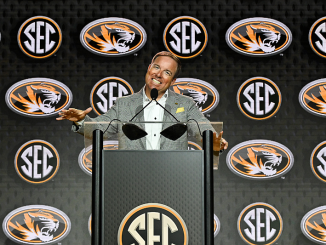 Man at a podium giving a speech at an SEC press conference with multiple SEC logos in the backdrop.
