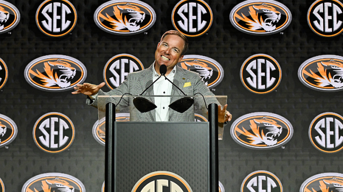 Man at a podium giving a speech at an SEC press conference with multiple SEC logos in the backdrop.