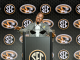 Man at a podium giving a speech at an SEC press conference with multiple SEC logos in the backdrop.