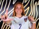Smiling female LSU soccer player wearing jersey number 2 holds up her jersey in front of a patterned wall, forming an L with her left hand.
