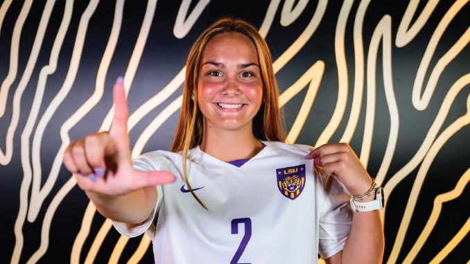 Smiling female LSU soccer player wearing jersey number 2 holds up her jersey in front of a patterned wall, forming an L with her left hand.