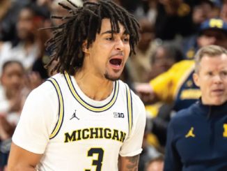 Michigan Wolverines basketball player with dreadlocks, number 3, shouting on the court; coach in blue and maize in the background.
