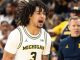 Michigan Wolverines basketball player with dreadlocks, number 3, shouting on the court; coach in blue and maize in the background.