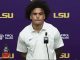 Athlete in a white LSU polo at a press conference, purple backdrop with LSU logos and a microphone in front.