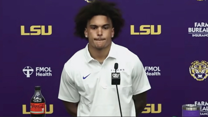 Athlete in a white LSU polo at a press conference, purple backdrop with LSU logos and a microphone in front.
