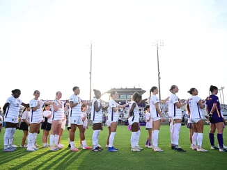 Line of female soccer players in white jerseys holding hands along the sideline on a sunny day at the stadium field.