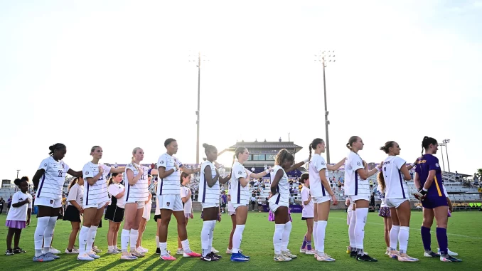 Line of female soccer players in white jerseys holding hands along the sideline on a sunny day at the stadium field.
