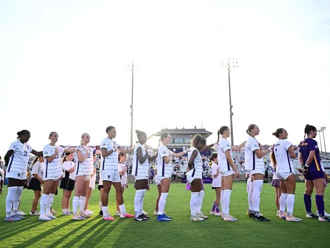 Line of female soccer players in white jerseys holding hands along the sideline on a sunny day at the stadium field.