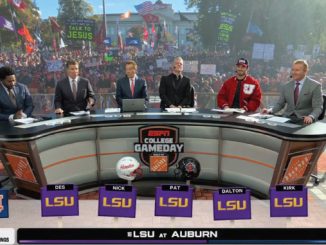 ESPN College GameDay broadcast on a sunlit campus plaza, hosts seated at a curved desk with LSU branding while a large crowd holds colorful signs in the background.