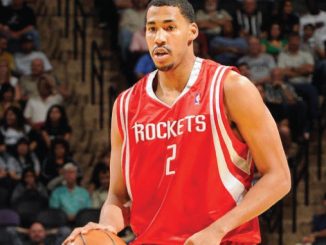 Basketball player in a red Houston Rockets jersey #2 dribbling the ball during a game with fans in the stands behind him.