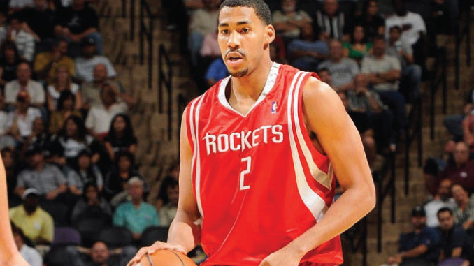 Basketball player in a red Houston Rockets jersey #2 dribbling the ball during a game with fans in the stands behind him.