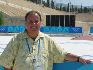 Smiling man in a yellow floral shirt standing by a pool at the Athens 2004 Olympic venue, wearing a blue lanyard and badge.