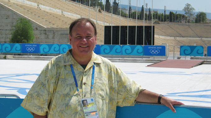 Smiling man in a yellow floral shirt standing by a pool at the Athens 2004 Olympic venue, wearing a blue lanyard and badge.