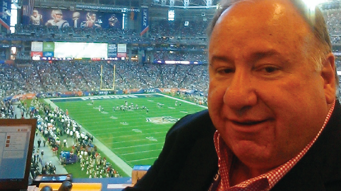 Man in a red checkered shirt in a stadium press box, overlooking a football game on the field outside the foreground.