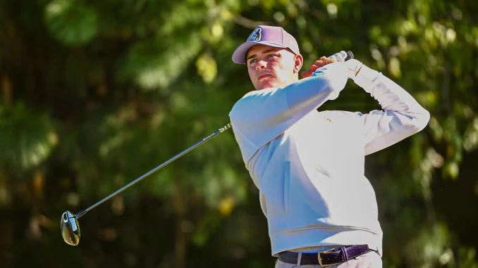 Male golfer in a light jacket and pink cap mid-swing on a sunny golf course outside the frame of the ball.