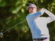 Male golfer in a light jacket and pink cap mid-swing on a sunny golf course outside the frame of the ball.