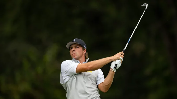 Male golfer in a gray polo and cap charges through a full swing with a driver on a golf course, focused on the ball area below him on the tee.