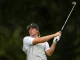 Male golfer in a gray polo and cap charges through a full swing with a driver on a golf course, focused on the ball area below him on the tee.