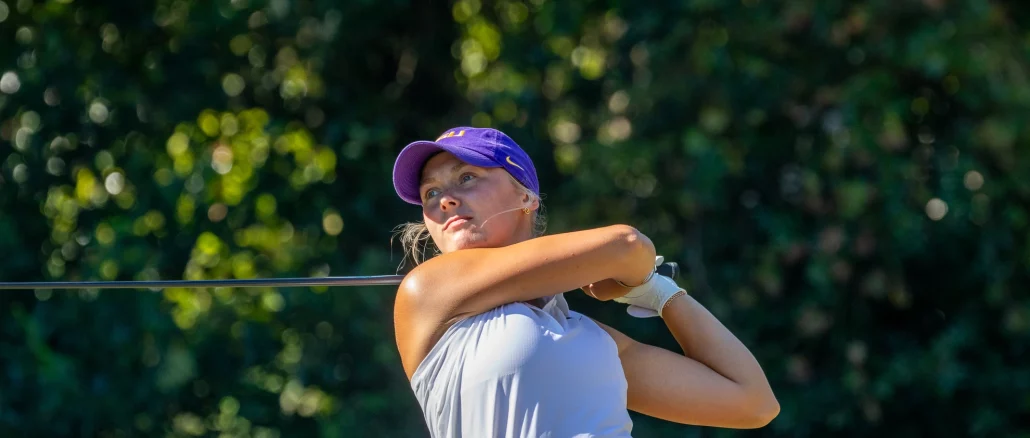 Female golfer in a purple cap and white top swings a golf club on a sunny course, with green trees in the background.