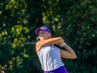 Female golfer in a purple cap and white top swings a golf club on a sunny course, with green trees in the background.