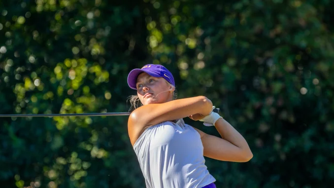 Female golfer in a purple cap and white top swings a golf club on a sunny course, with green trees in the background.