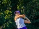Female golfer in a purple cap and white top swings a golf club on a sunny course, with green trees in the background.