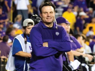 Smiling coach in a purple polo with arms crossed, standing on the sideline as photographers and a crowded stadium backdrop watch.