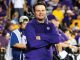 Smiling coach in a purple polo with arms crossed, standing on the sideline as photographers and a crowded stadium backdrop watch.