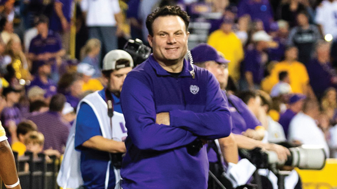 Smiling coach in a purple polo with arms crossed, standing on the sideline as photographers and a crowded stadium backdrop watch.