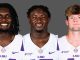 Three LSU football players smiling, wearing white jerseys with purple LSU logos, against a gray backdrop.