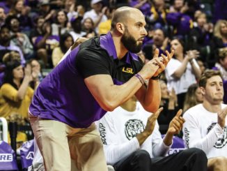 LSU coach in purple and gold leaning forward, clapping during a game with the crowd behind him.