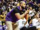 LSU coach in purple and gold leaning forward, clapping during a game with the crowd behind him.