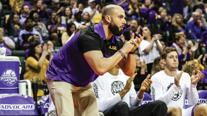 LSU coach in purple and gold leaning forward, clapping during a game with the crowd behind him.