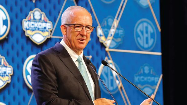 Man in a dark suit and teal tie speaks at a podium during an SEC kickoff event, blue patterned backdrop behind him.